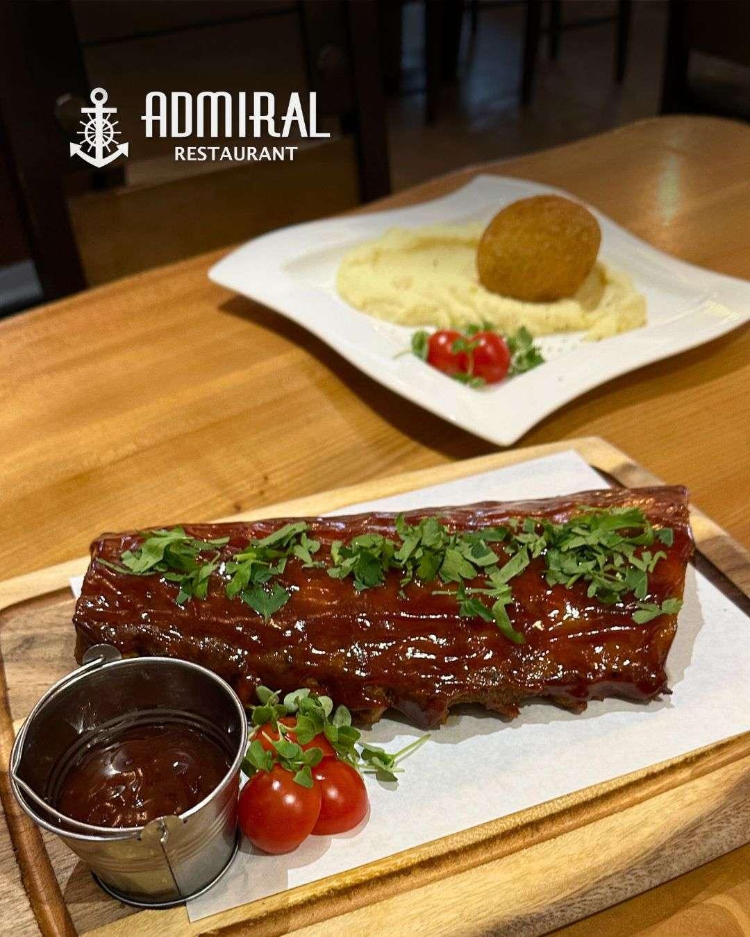 Barbecued pork ribs glazed in sauce and topped with chopped herbs, served with cherry tomatoes and dipping sauce on a wooden board. In the background, a croquette with mashed potatoes and garnish is on a white plate.
