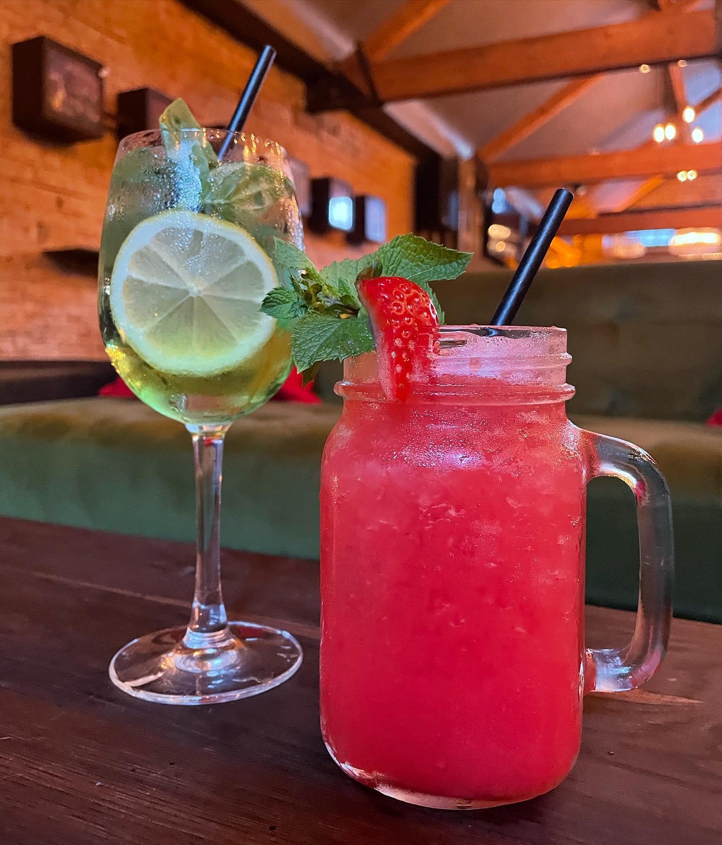 Two colorful cocktails sit on a dark wooden table in a cozy bar with warm lighting and exposed brick walls. The drink on the left is served in a wine glass, garnished with lemon slices and mint leaves, and has a clear, refreshing appearance. The drink on the right is a vibrant red beverage served in a mason jar with crushed ice, garnished with a strawberry slice and a sprig of mint, with a black straw in both drinks. The background shows a comfortable green sofa and wooden beams on the ceiling.