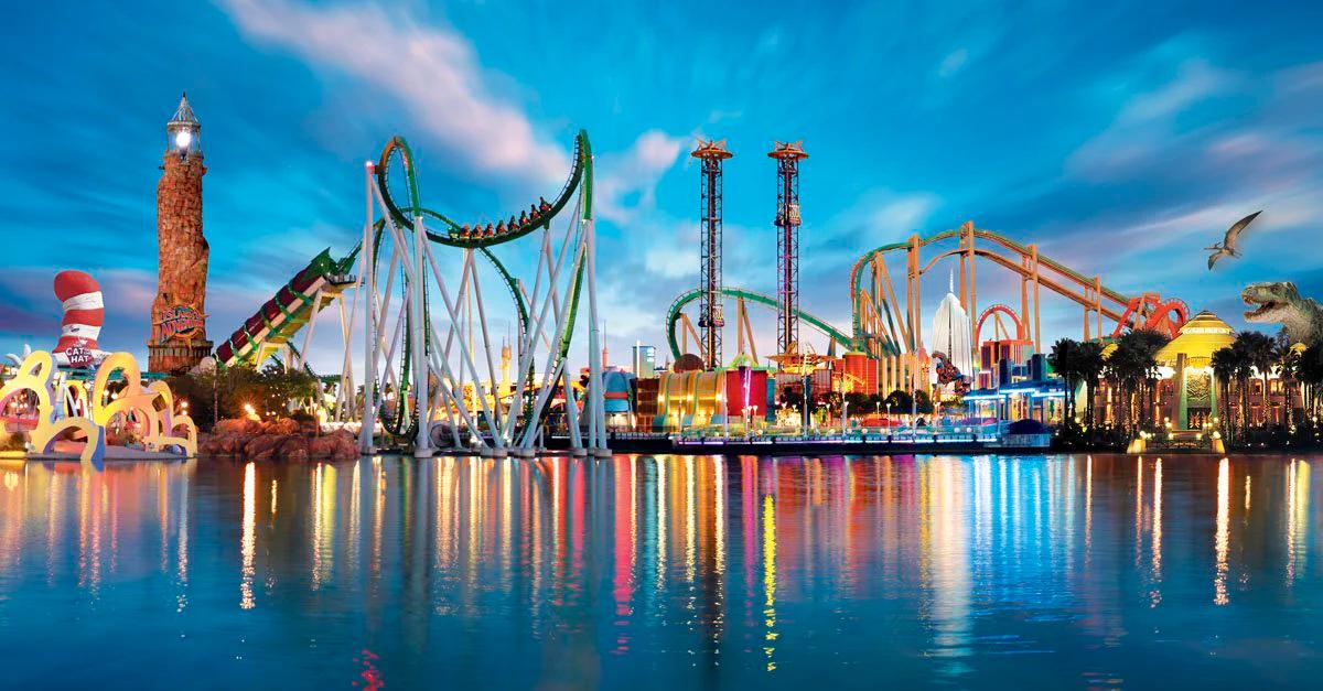 A colorful theme park with large roller coasters, tall drop towers, and various attractions reflected in a calm body of water, under a vibrant sky at dusk.