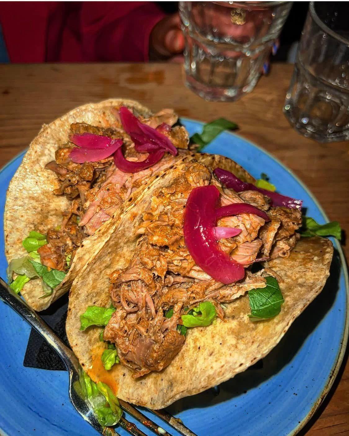 This image shows two tacos served on a blue plate. The tacos are filled with shredded, seasoned meat, topped with pickled red onions and some fresh greens, likely lettuce and spinach. A fork is resting on the plate, and in the background, there are two empty glasses and a person's hand. The setting suggests a casual dining experience. The tacos look hearty and flavorful!