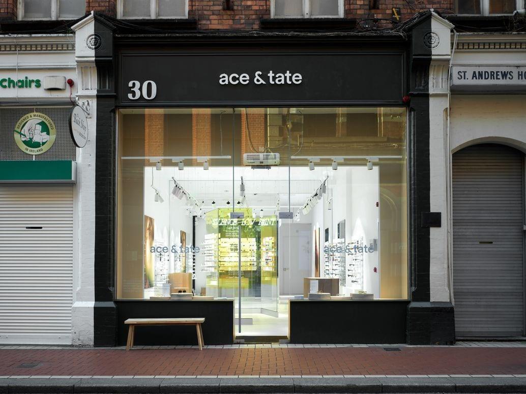 Front view of the ace & tate eyeglasses store at 30 St. Andrews. The storefront features large clear glass windows and doors revealing a bright interior with eyeglass displays along the walls. A small wooden bench is positioned outside on the sidewalk. The building facade is black and white with brickwork above.