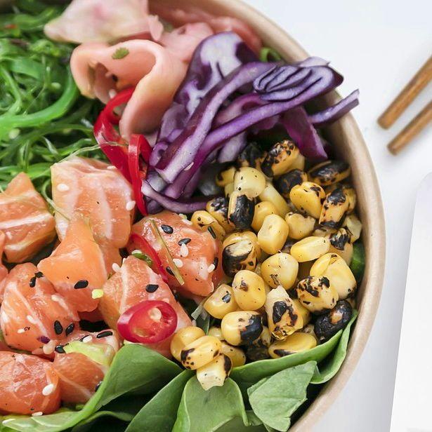 Fresh poké bowl featuring raw salmon cubes, charred corn, sliced red cabbage, seaweed salad, pickled ginger, red chili, and leafy greens in a round bowl.