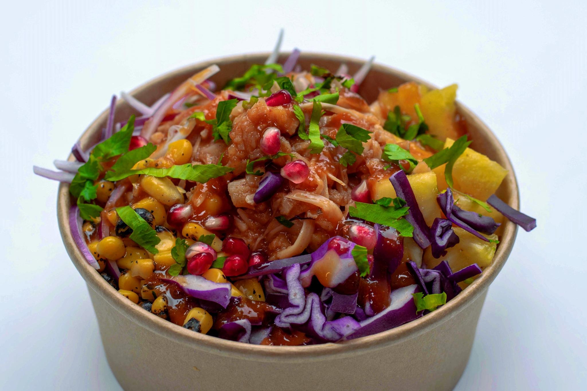 Colorful Hawaiian poké bowl filled with marinated fish, purple cabbage, corn, pineapple, pomegranate seeds, red onion, and fresh herbs in a brown compostable container.