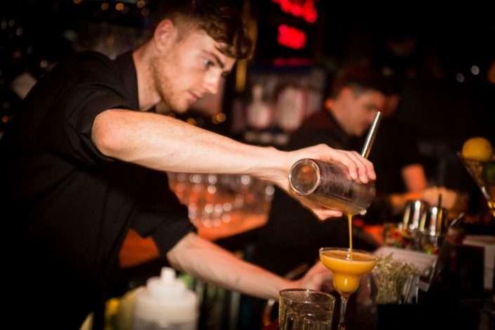A bartender in a dimly lit bar is pouring a bright orange cocktail from a shaker into a martini glass. He is wearing a black shirt and is focused on the drink. The bar counter is lined with various glasses and garnishes, and another bartender is working in the background.