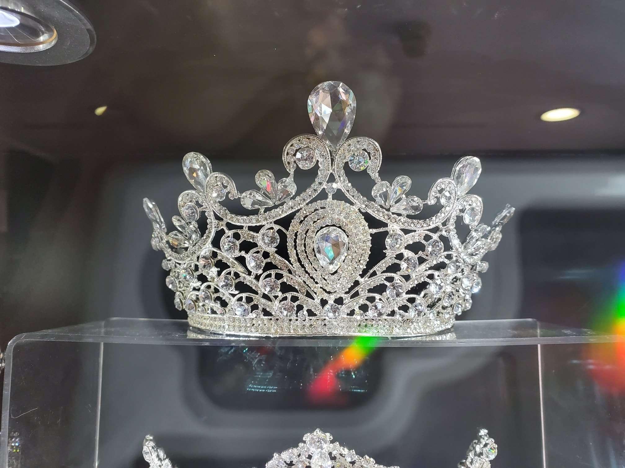 A luxurious silver tiara adorned with large teardrop and round-cut clear gemstones. The intricate filigree design features swirls, loops, and floral accents, creating an elegant and regal appearance. The tiara is displayed on a transparent stand under soft lighting, with rainbow light reflections visible below.