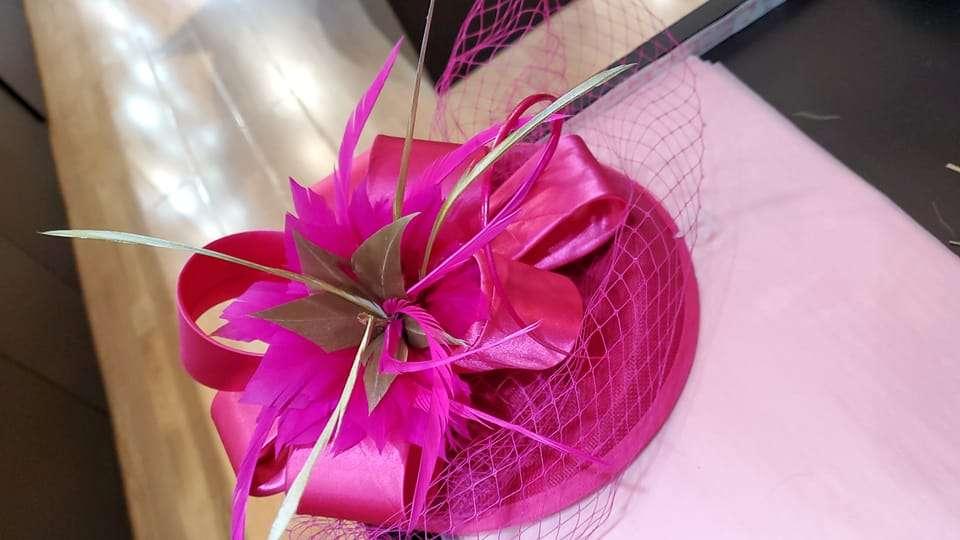 A vibrant fuchsia pink fascinator hat featuring a large satin ribbon, bright pink feathers, and light green decorative spikes, topped with a delicate mesh veil. The hat is placed on a light pink fabric surface next to a wooden floor.