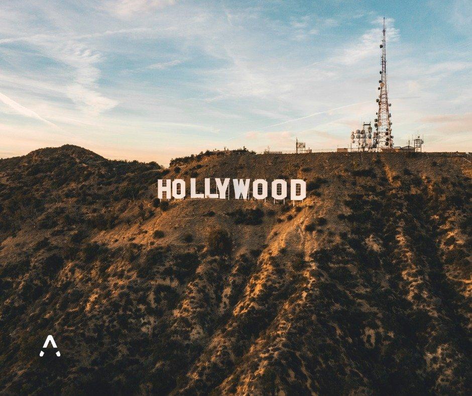 Aerial view of the iconic white Hollywood sign on a rugged hillside at sunset, with a communications tower visible nearby under a blue sky with soft clouds.