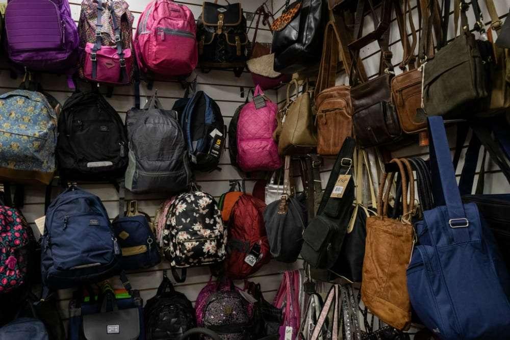 A store wall display featuring a variety of backpacks and handbags, including floral, solid, and leather styles in colors like pink, black, brown, and blue.
