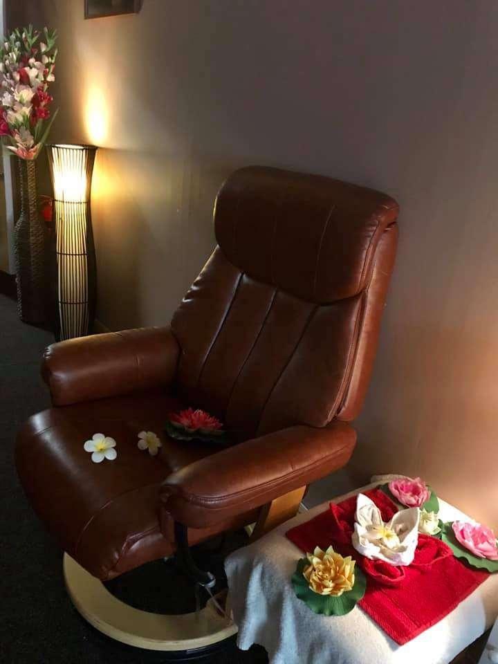 A brown leather recliner chair with decorative flowers on it, next to a small table with a red towel and more flowers. A tall lamp and flower vase provide soft lighting in the background.