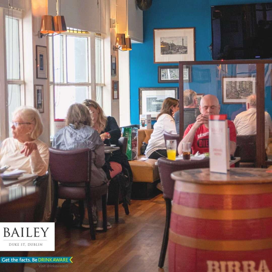 This image shows the cozy interior of Bailey Bar & Café. Diners sit at tables enjoying drinks and meals, surrounded by warm lighting, wooden floors, and framed black-and-white photos on blue walls.