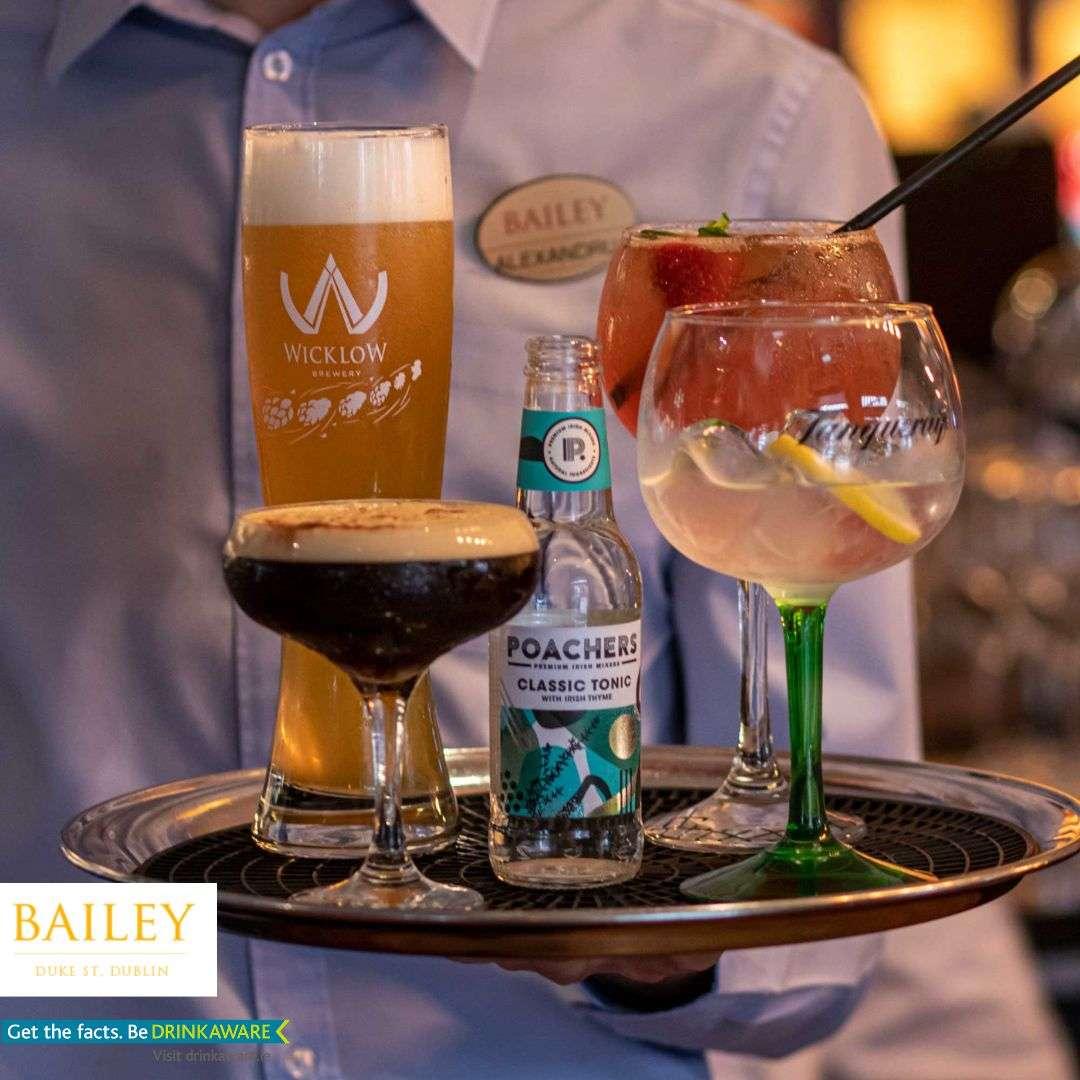 This image features a server at **Bailey Bar & Café** carrying a tray of drinks, highlighting a vibrant and upscale bar experience. The tray includes a tall glass of **Wicklow Brewery beer**, an **espresso martini**, a bottle of **Poachers Classic Tonic**, and two stylish cocktails — one garnished with **strawberries and mint**, and the other with a **lemon twist**. The drinks are elegantly presented, emphasizing the variety of options and the attention to detail typical of a well-established bar in **Duke Street, Dublin**. The Bailey logo and “Be Drinkaware” reminder also appear, reinforcing a responsible yet refined drinking atmosphere.