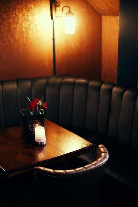A dimly lit, cozy booth in a lounge or bar with dark leather seating surrounding a wooden table. The table is decorated with a small vase of red flowers and a glowing candle, creating a warm and intimate ambiance. A wall sconce above casts soft light on the textured walls.
