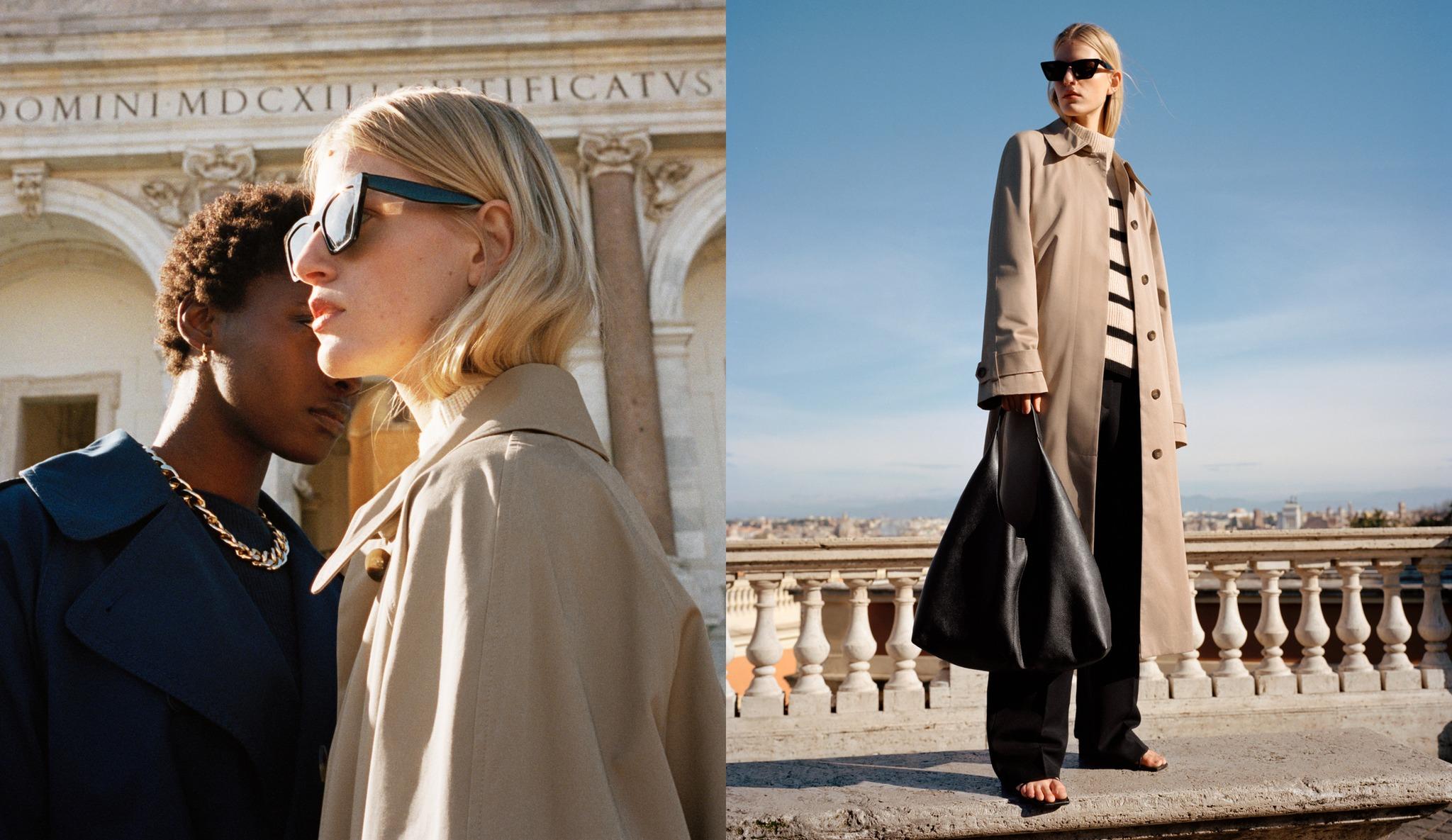 Two fashion-forward women in trench coats against a classical architectural backdrop. On the left, a close-up of two models—one in a navy coat with a gold chain necklace, the other in a beige trench and dark sunglasses—stand in soft sunlight near a historic stone building. On the right, the woman in the beige trench coat is seen full-length, wearing black pants, a striped sweater, and holding a large black slouchy bag, posing confidently on a terrace overlooking a city skyline under a clear blue sky.