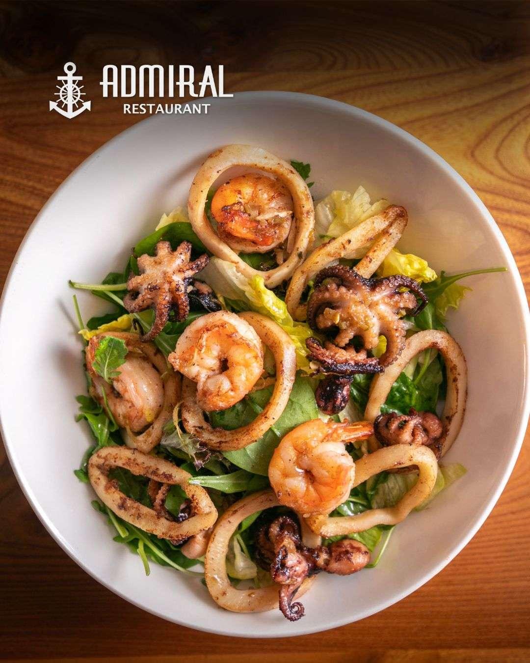 Bowl of mixed green salad topped with grilled prawns, calamari rings, and baby octopus, served on a wooden table under natural light.