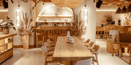 Warmly lit restaurant interior featuring a long communal wooden table with chairs, a bar with stools, and large planters filled with dried grasses.