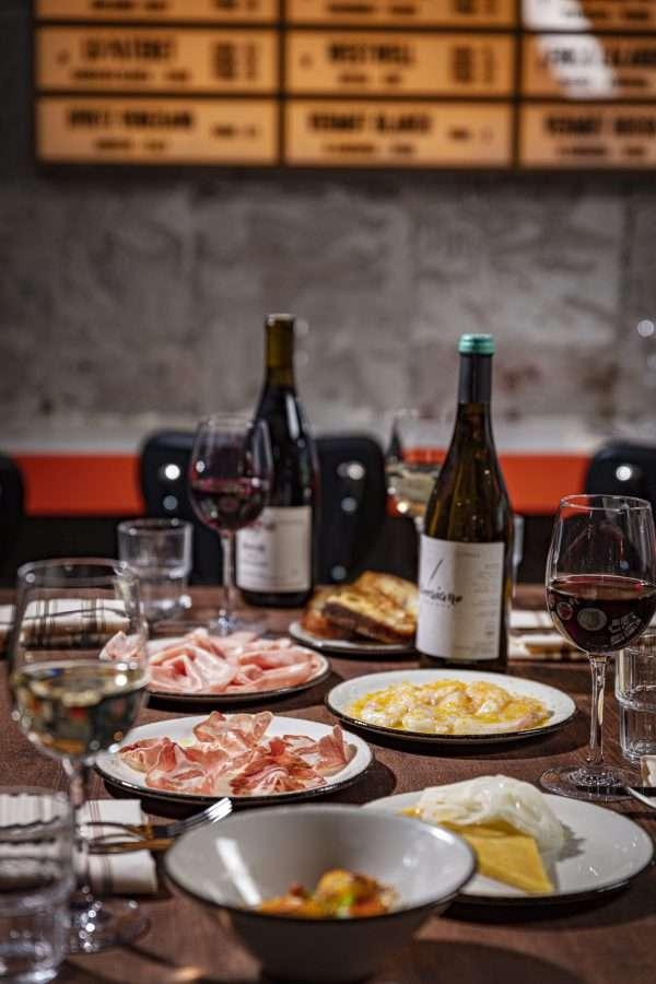 A rustic dining table set with several glasses of red and white wine, bottles, and plates of charcuterie, cheese, and other appetizers under a softly lit wine menu on the wall.
