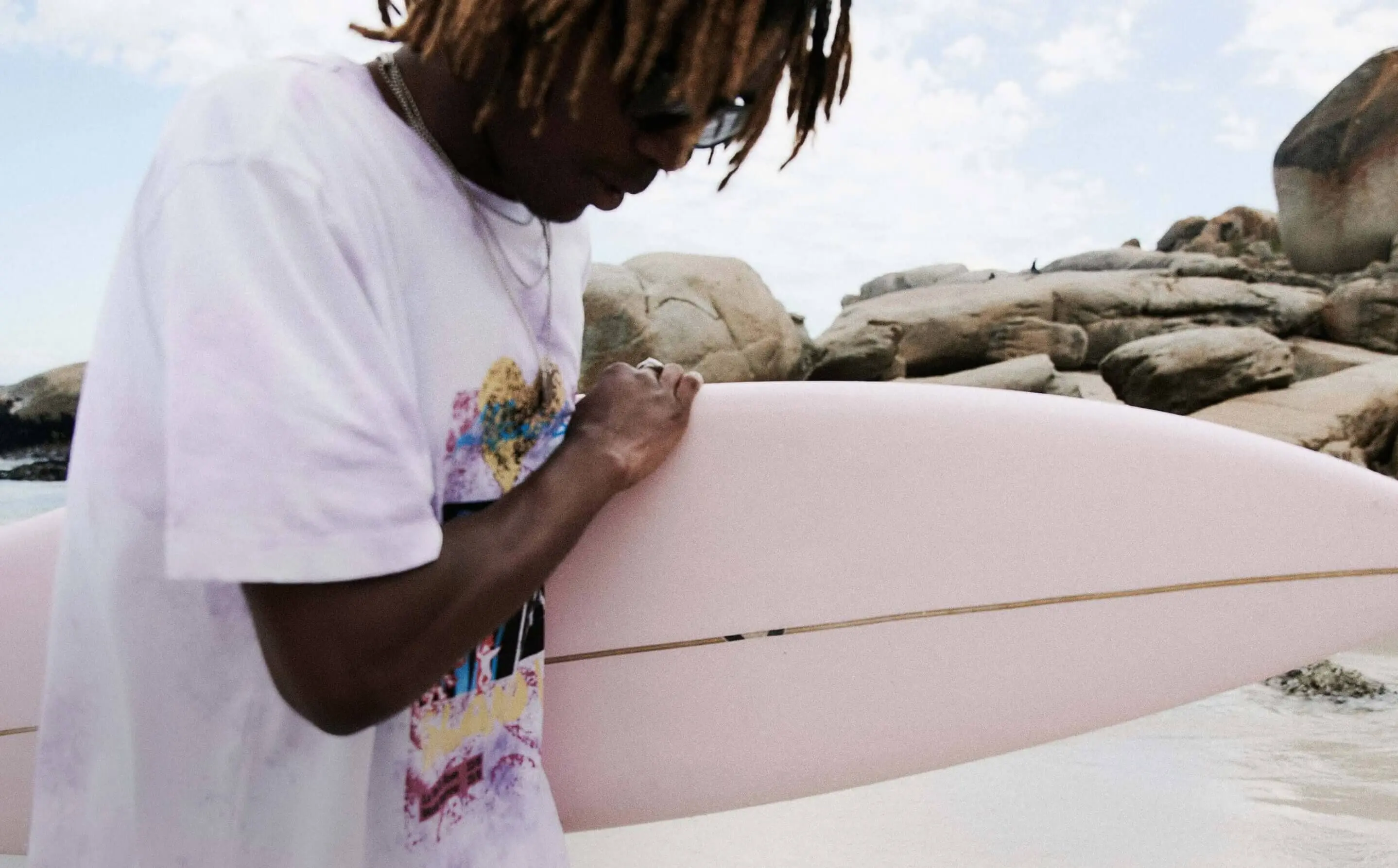 A person with dreadlocks wearing a white graphic T-shirt and sunglasses holds a pale pink surfboard near rocky shore and ocean water.