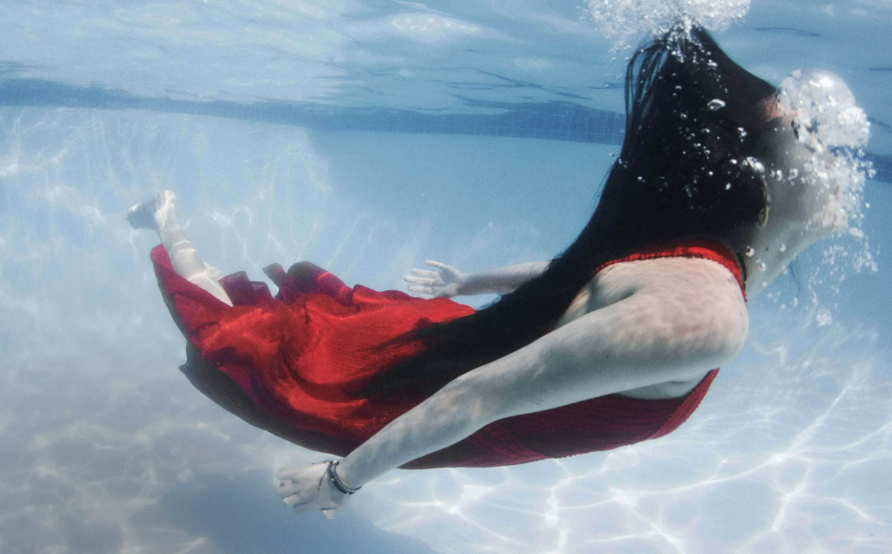 Underwater shot of a person swimming horizontally. They have long black hair flowing behind them and are wearing a flowing red dress. Bubbles are visible around their head, and the pool floor and walls are faintly visible through the clear water.