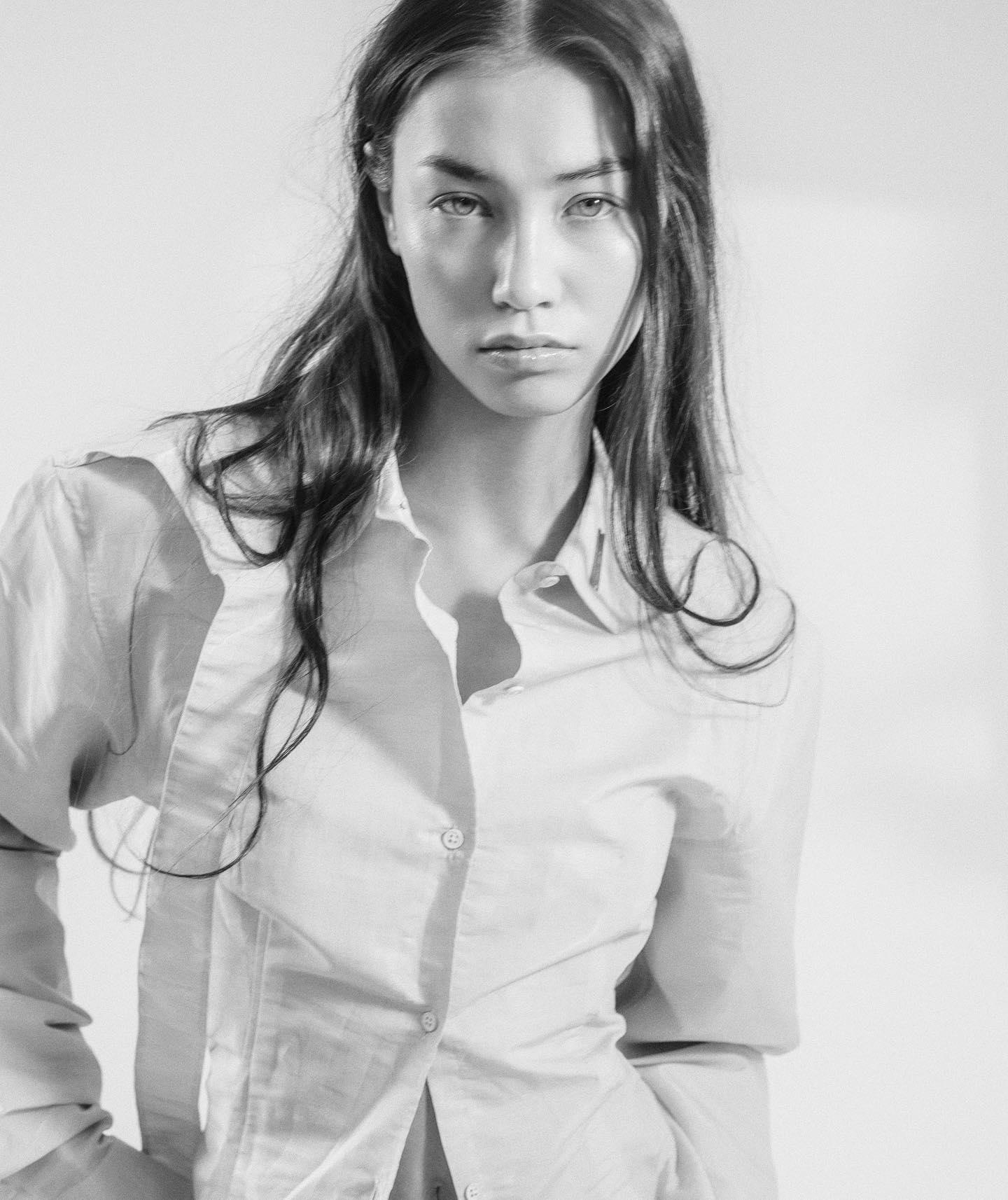 Black and white portrait of a woman with long, dark hair wearing a button-up shirt. She gazes directly at the camera with a serious expression, her hair slightly tousled, and soft shadows falling on the wall behind her.