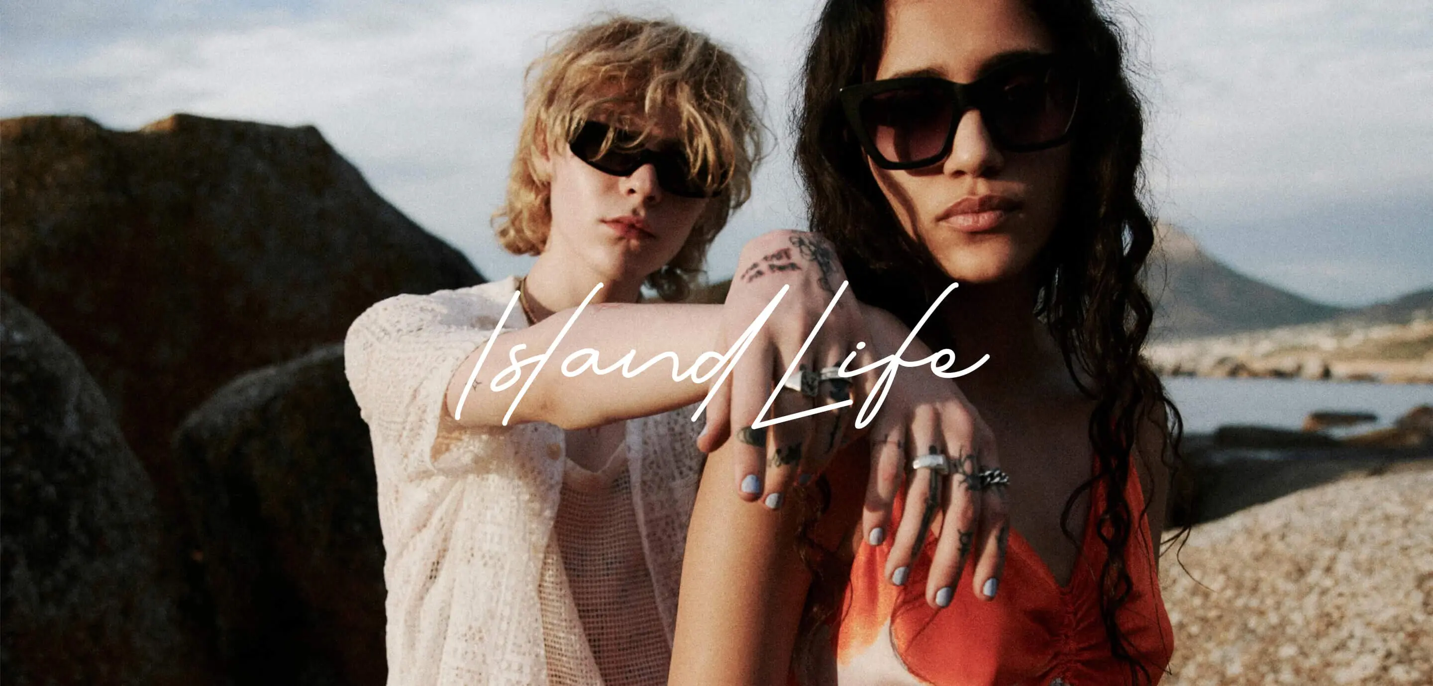 Two young women wearing sunglasses pose outdoors near rocky terrain by the water. One has short blonde hair and a white crochet top; the other has long dark hair and an orange top. The text 'Island Life' overlays the image.