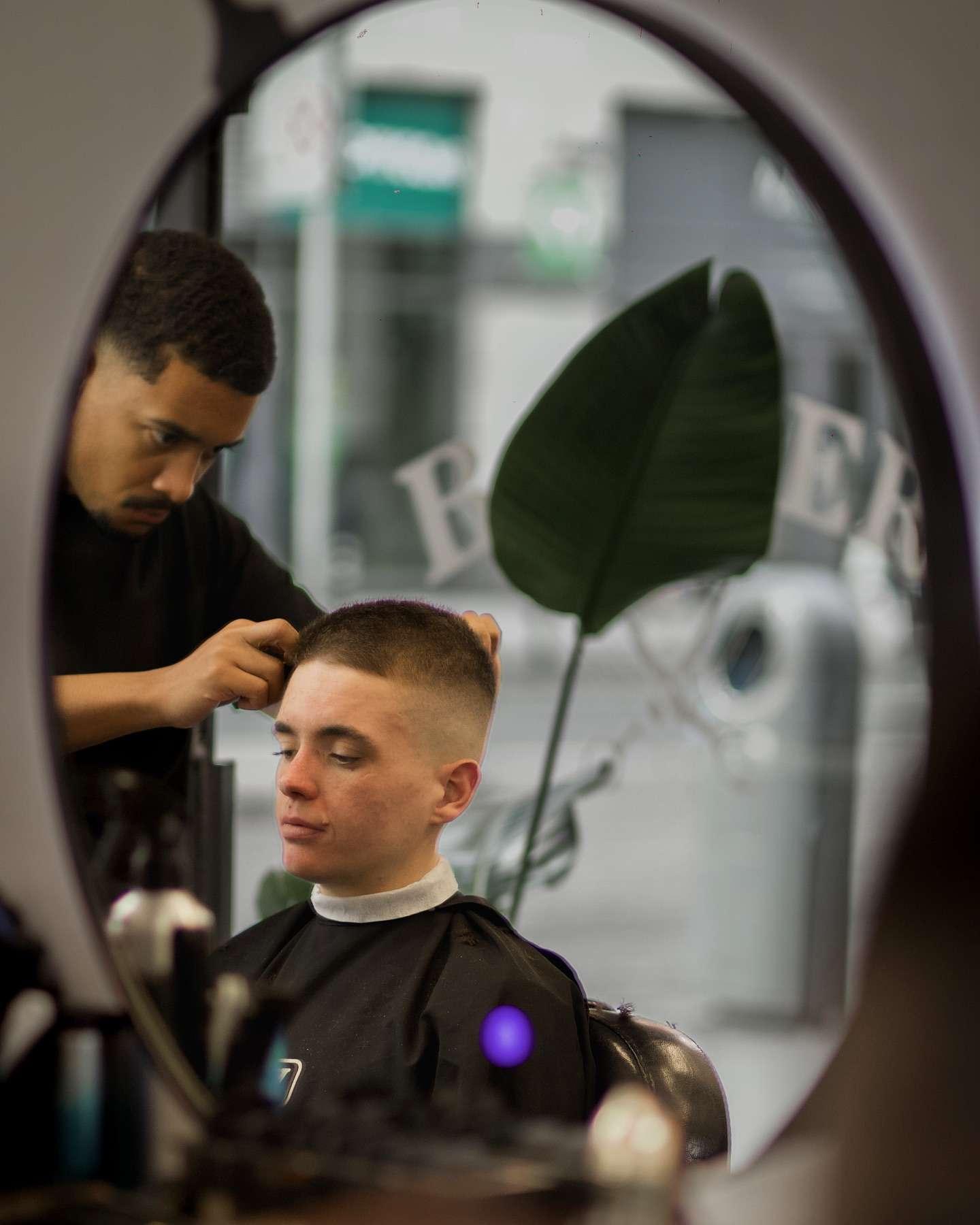 Reflection in a round mirror showing a barber giving a young man a buzz cut with a high skin fade, as the client sits calmly in the barber chair inside a stylish barbershop.