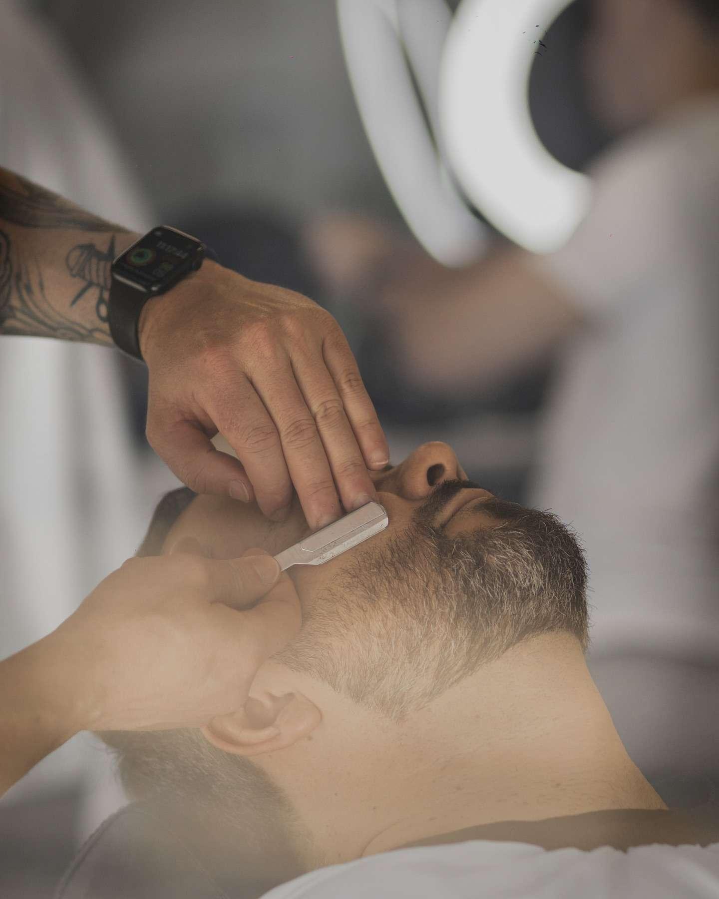 Barber shaping a man's beard with a straight razor while the client reclines in the chair.