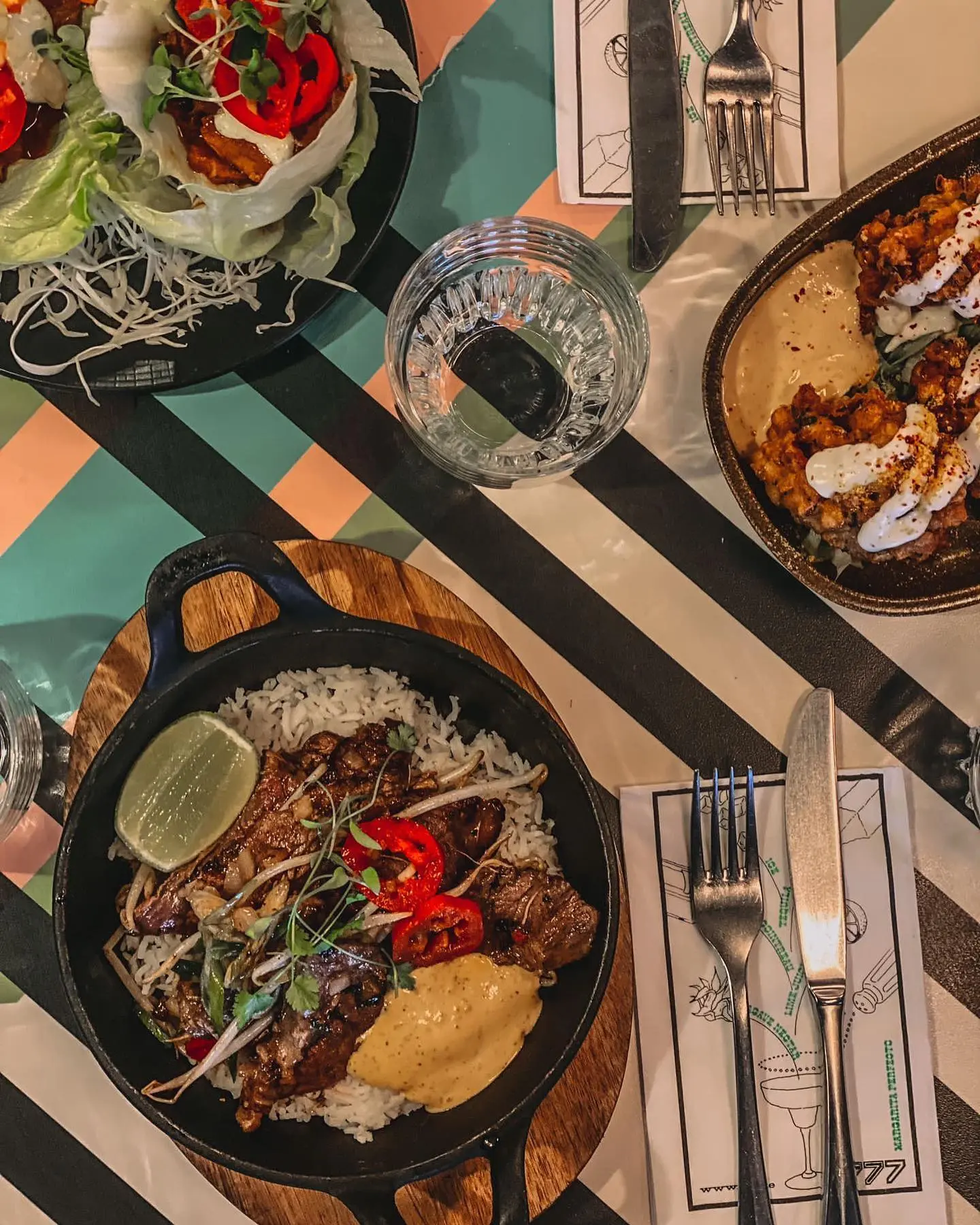 An overhead view of a colorful table set with three modern Mexican dishes. At the bottom center, a cast iron skillet on a wooden board contains grilled beef slices over white rice, topped with red chili slices, microgreens, a dollop of creamy yellow sauce, and a lime wedge. To the top left, lettuce cups filled with shredded vegetables and spicy toppings are arranged on a black plate. At the top right, a brown dish holds crispy cauliflower bites drizzled with white sauce and served with a beige dip.