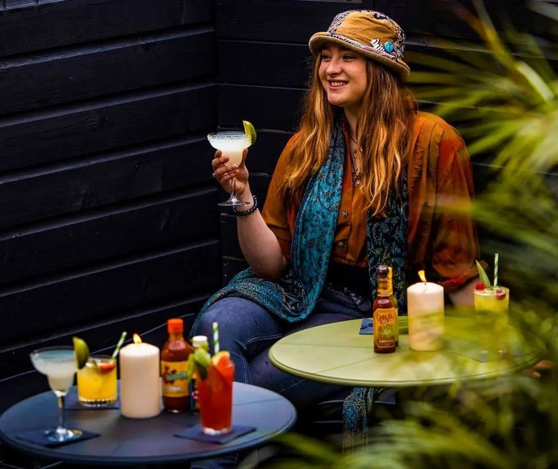 A woman sitting outdoors at a small table, smiling and holding a margarita garnished with lime. She is wearing a brown hat with a turquoise brooch, a rust-colored shirt, and a blue patterned scarf. The table is set with several colorful cocktails, bottles of hot sauce, and lit candles, creating a cozy and vibrant atmosphere.