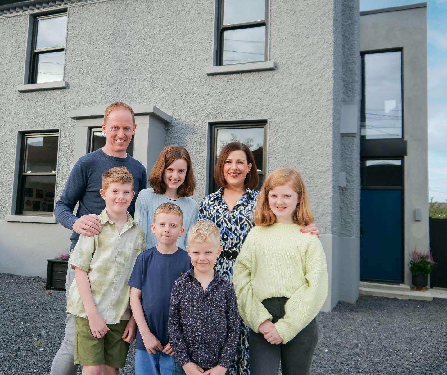 A family of seven—two adults and five children—posing and smiling in front of a modern grey house with black-trimmed windows and a blue front door. The adults stand behind the children, who are dressed casually and standing on a gravel driveway. The setting is bright and cheerful. Ask ChatGPT