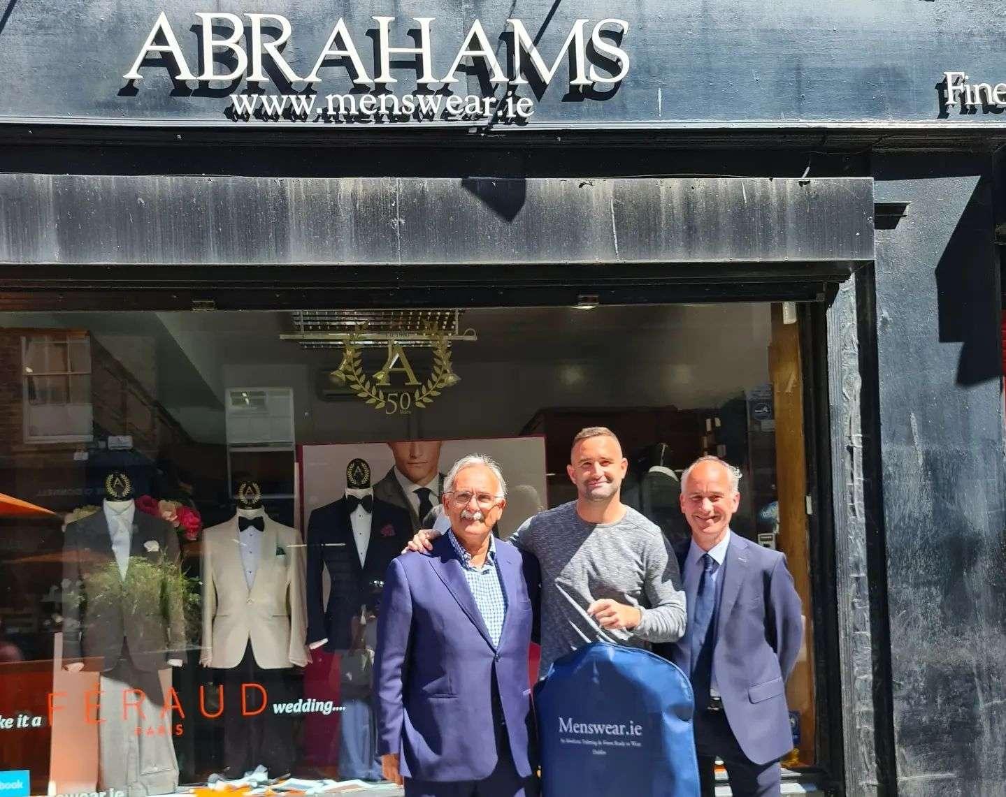 Three men standing in front of a menswear shop named "Abrahams" with the website menswear.ie visible on the sign. The men are smiling, two in suits and one in casual clothing holding a blue garment bag with the same website printed on it. Behind them, mannequins dressed in formal suits are displayed in the shop window.