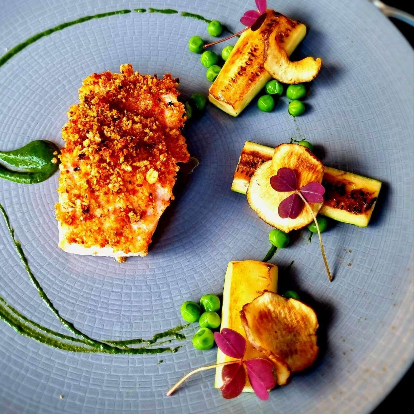 A gourmet dish artfully plated on a textured gray plate. On the left side, there is a piece of crusted salmon with a golden crumb topping. On the right side, there are three small portions of grilled vegetables, possibly zucchini, garnished with green peas and thin slices of roasted root vegetables topped with small purple edible leaves. A swirl of green sauce decorates the plate, adding a vibrant touch.