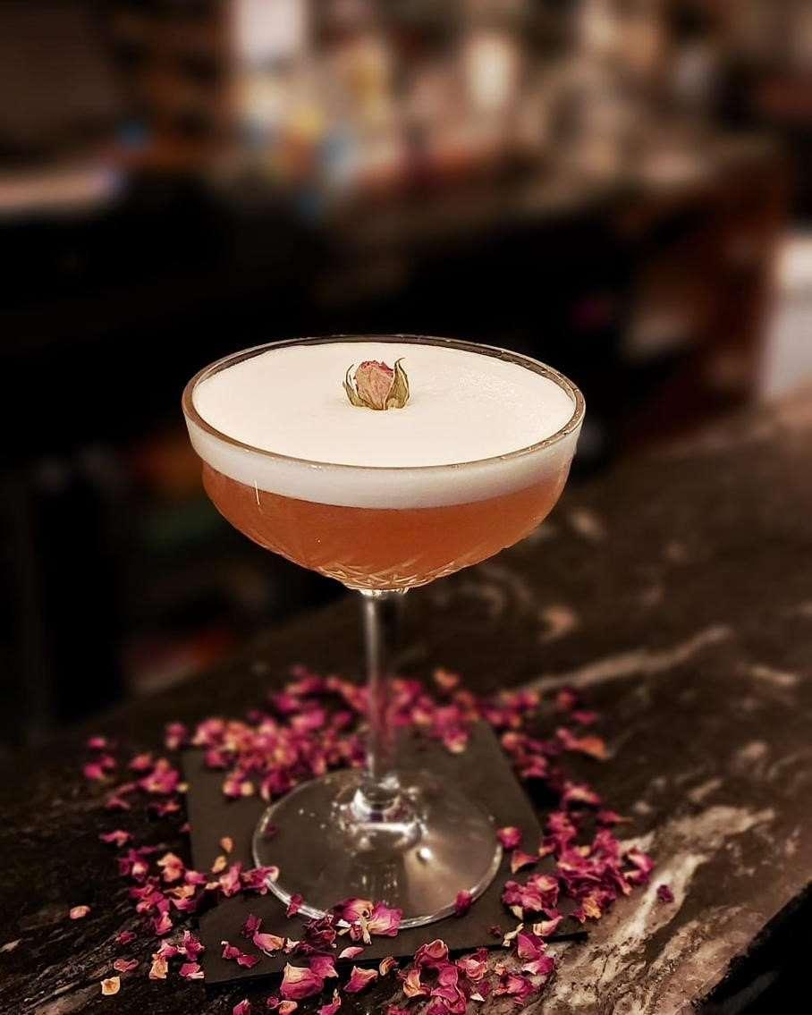 A coupe glass with a frothy light pink cocktail topped with a small dried rosebud garnish. The glass is placed on a dark marble bar surface scattered with dried pink rose petals. The background is softly blurred, focusing attention on the drink.