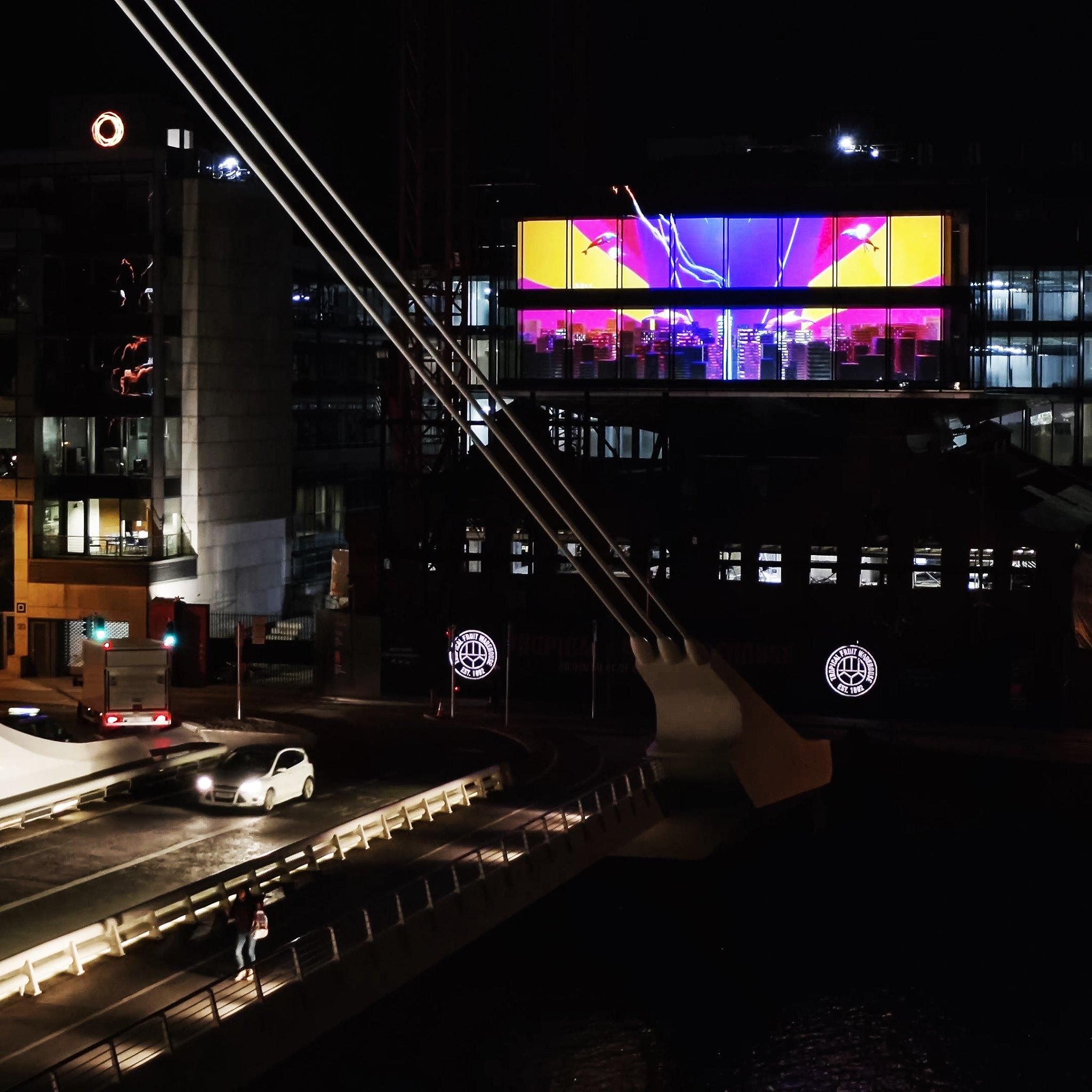 Night view of a modern bridge with white cables and light trails from cars, featuring a large building in the background displaying a vibrant digital art installation in purple, yellow, and pink tones.
