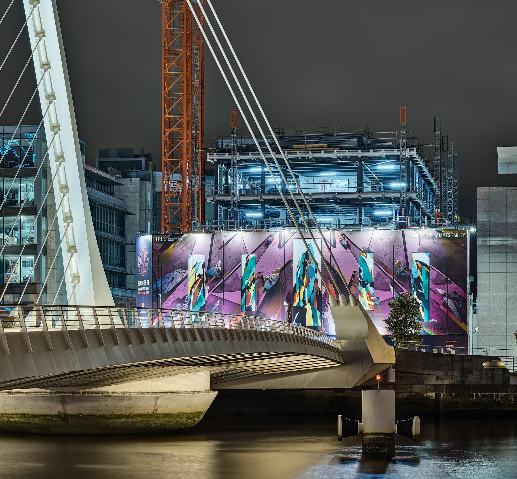 Illuminated modern bridge leading to a construction site covered in a large, colorful geometric mural featuring abstract figures, with a steel frame building and orange crane in the background.