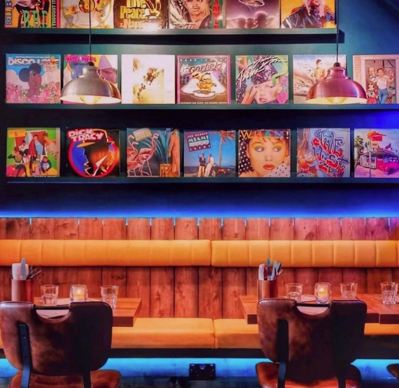 A cozy restaurant interior featuring wooden tables and leather chairs with mustard-yellow cushioned bench seating. The wall above the seating is decorated with colorful retro album covers displayed on shelves. Two hanging pendant lights with metal shades provide warm lighting, and blue LED strip lights run along the bottom of the wall.