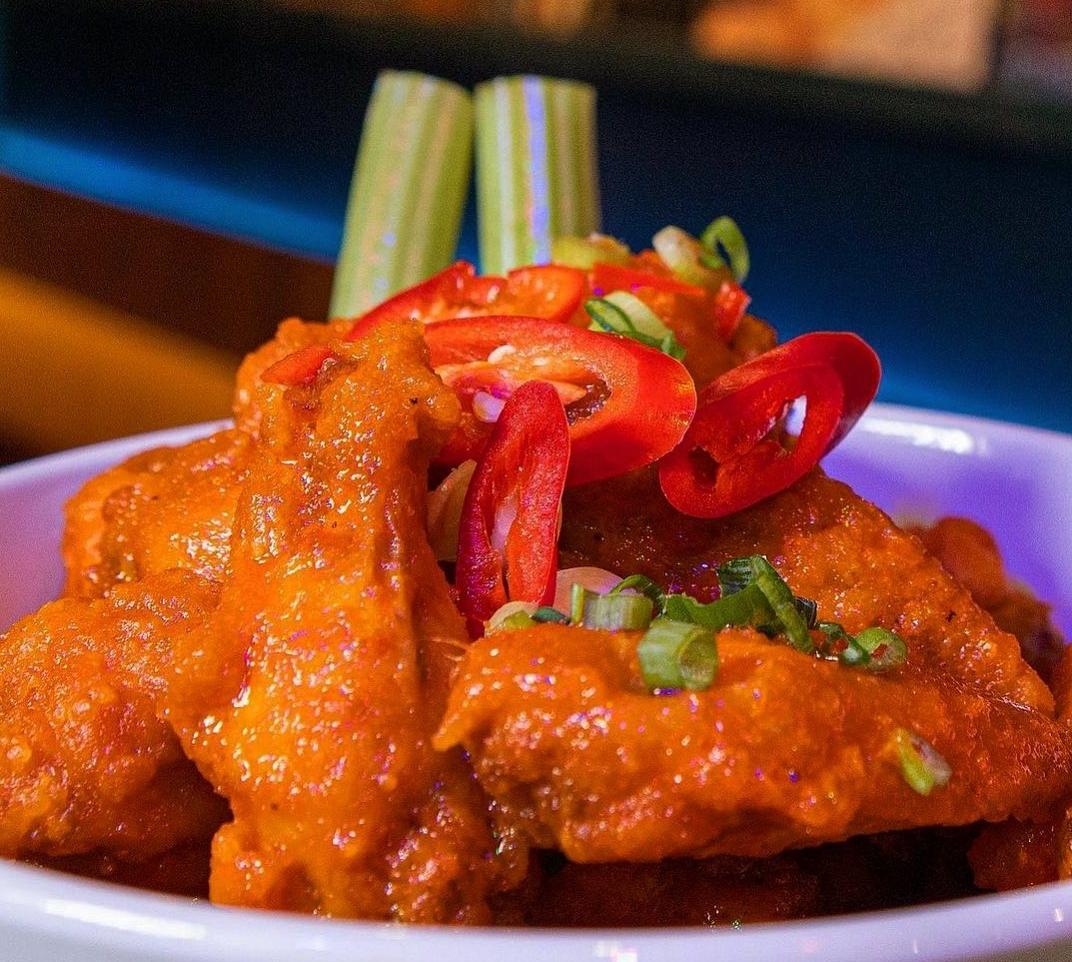 Close-up of a bowl of spicy buffalo chicken wings garnished with sliced red chili peppers, chopped green onions, and two celery sticks.