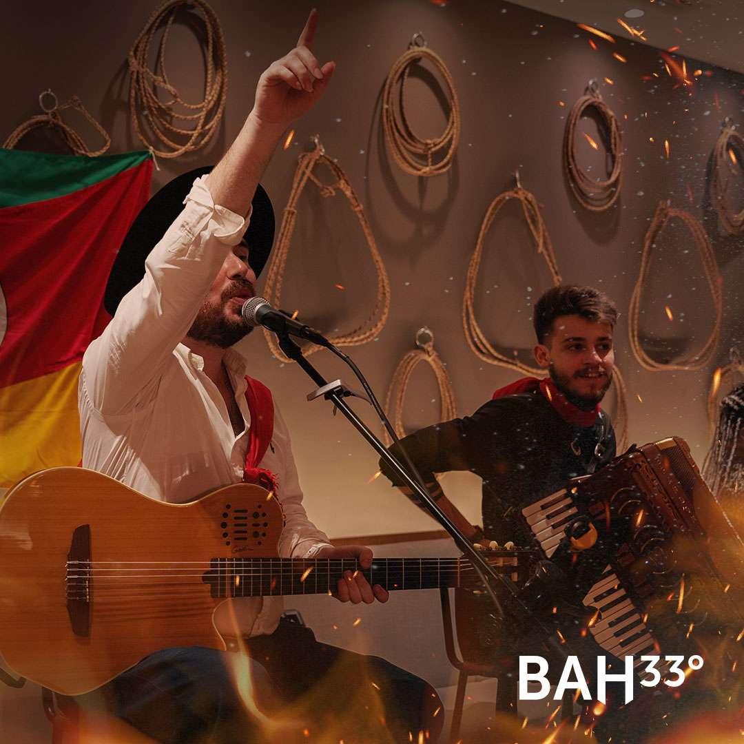 A traditional live music performance featuring a man singing and playing guitar while another musician plays accordion. Both are dressed in regional attire, performing in front of a wall decorated with coiled ropes and a red, green, and yellow flag, with warm lighting and sparks enhancing the festive atmosphere.