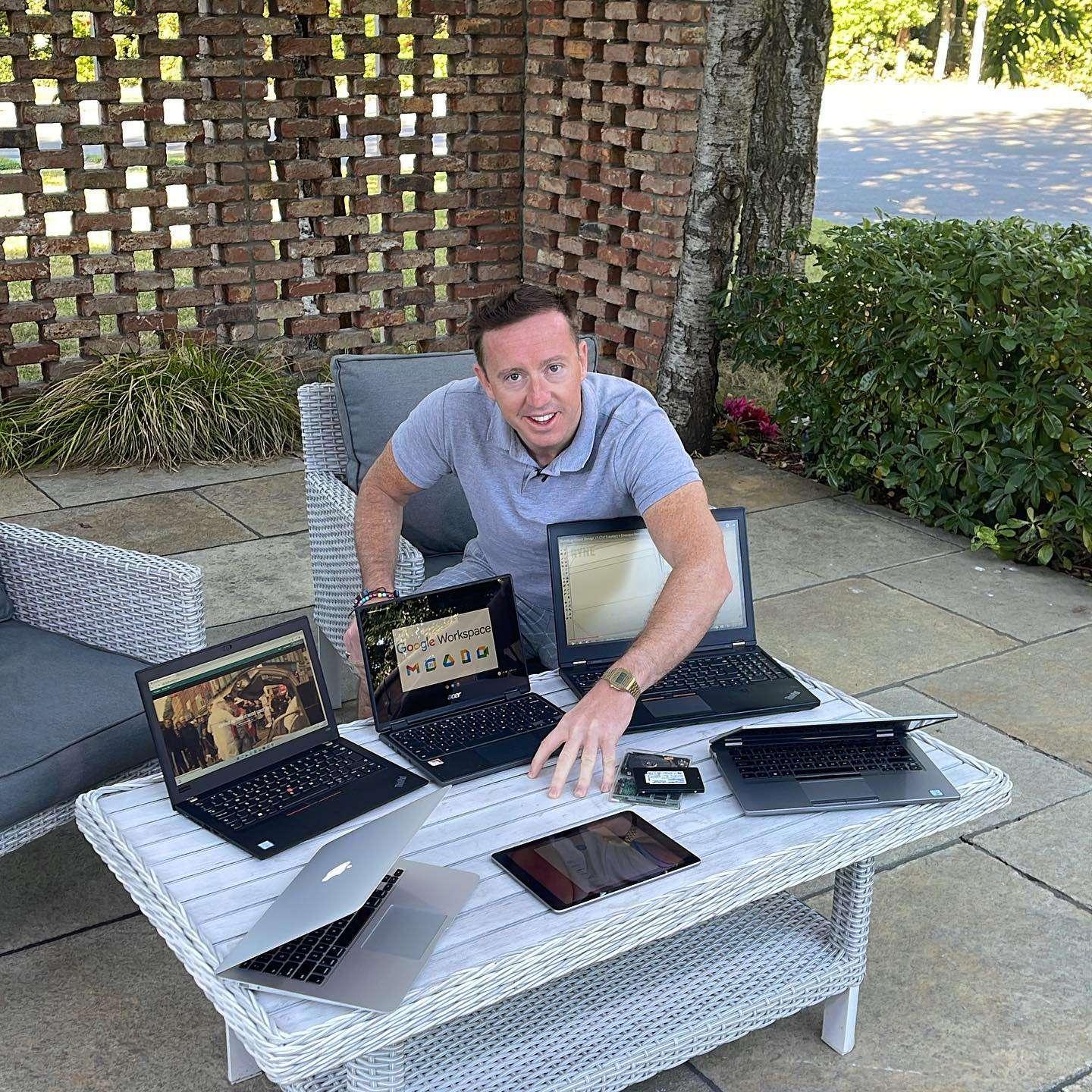 A man sitting outdoors at a white wicker table surrounded by several open laptops and a tablet, appearing to work on or display various devices. The setting includes patio furniture, a brick privacy wall, and greenery in the background.