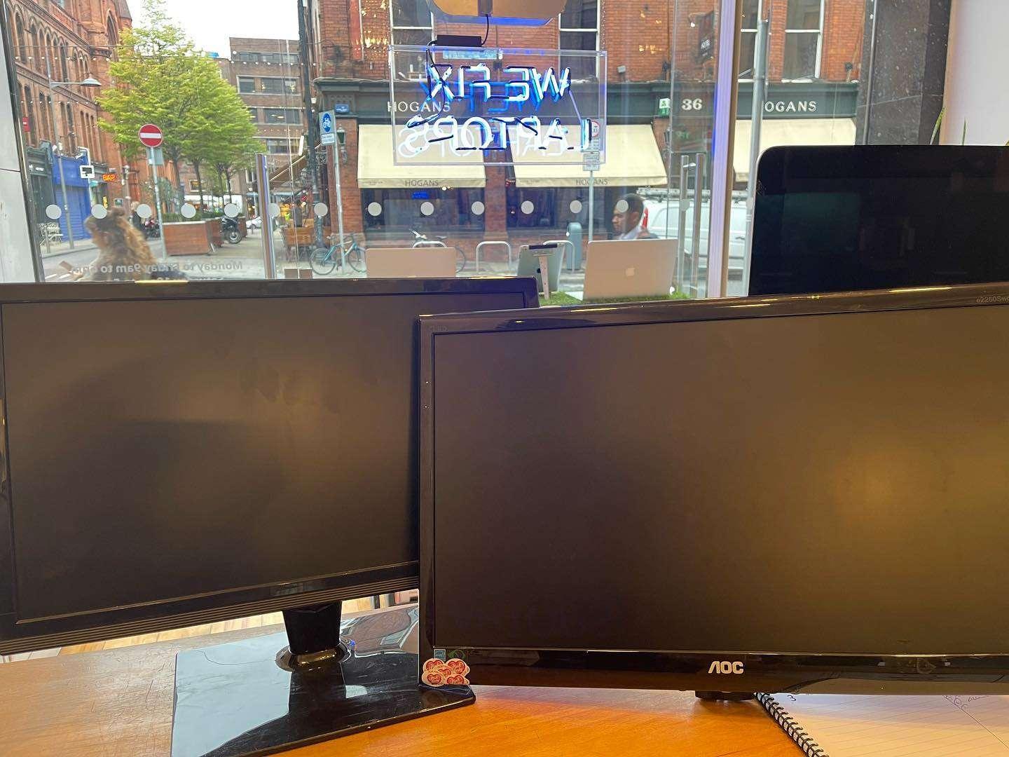 Two blank computer monitors on a desk in front of a large window, with a view of a city street, including a neon sign with mirrored text and a café named Hogans across the road.