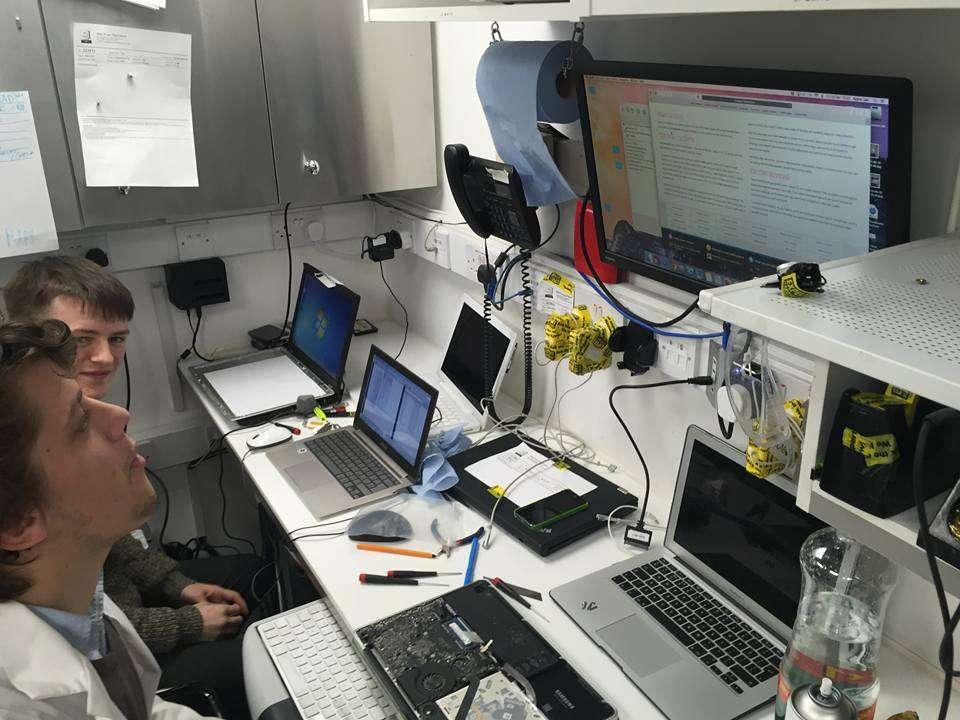 Two men sit at a cluttered electronics workbench with several open laptops, tools, and a disassembled laptop. A large monitor on the wall displays technical documentation, and various cables and tape are organized on shelves.