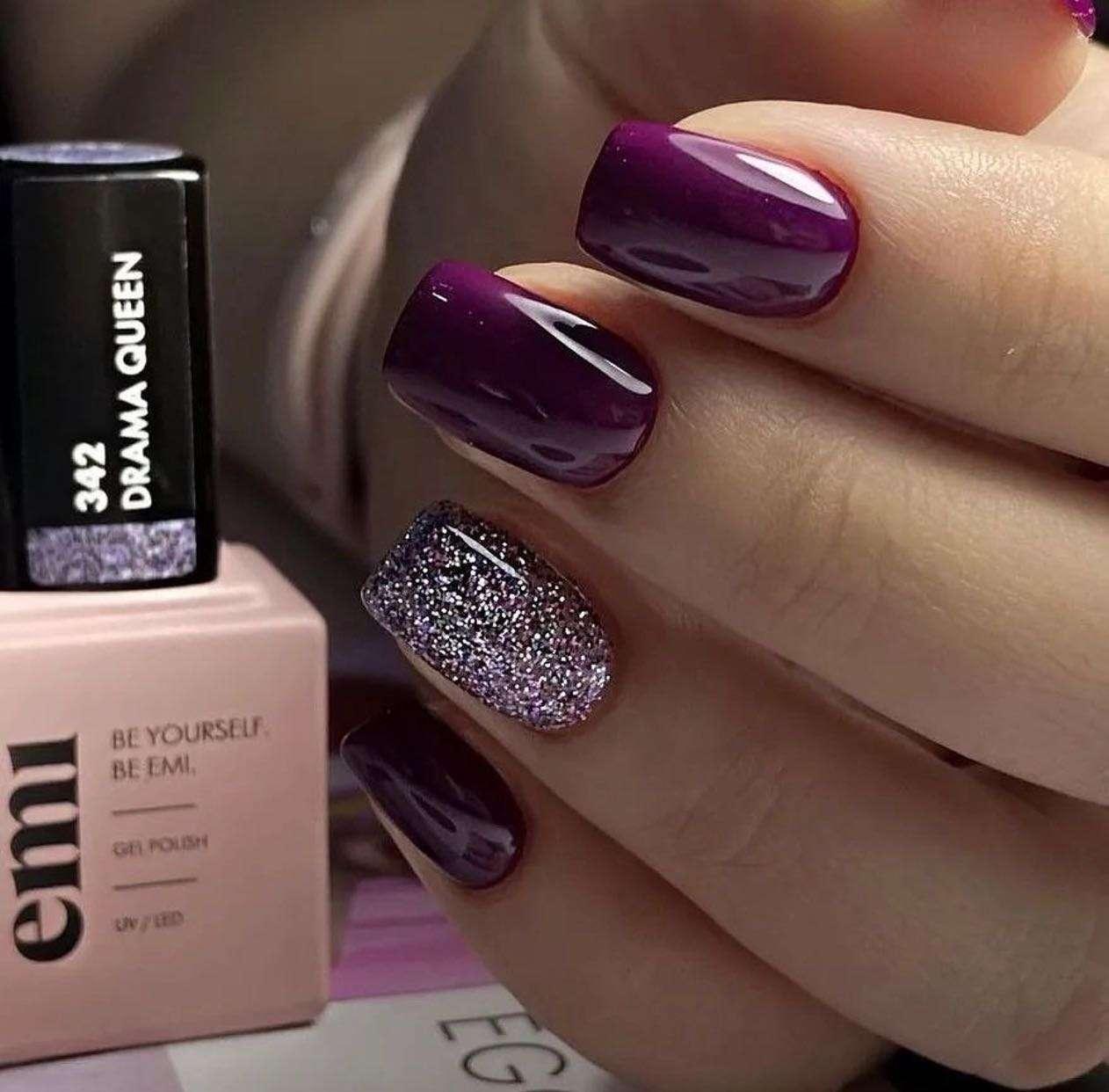 Close-up of a hand showing freshly manicured nails. Four nails are painted glossy dark purple, and the ring finger nail has purple glitter polish. Next to the hand is a bottle of EMI gel polish labeled "342 DRAMA QUEEN."
