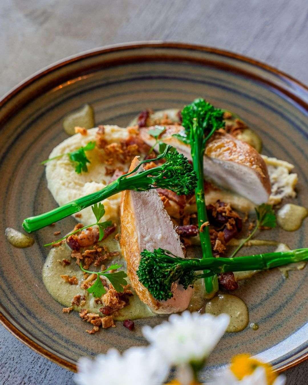 Sliced roasted chicken breast served with mashed potatoes, tenderstem broccoli, crispy bits, and a light green sauce on a ceramic plate.