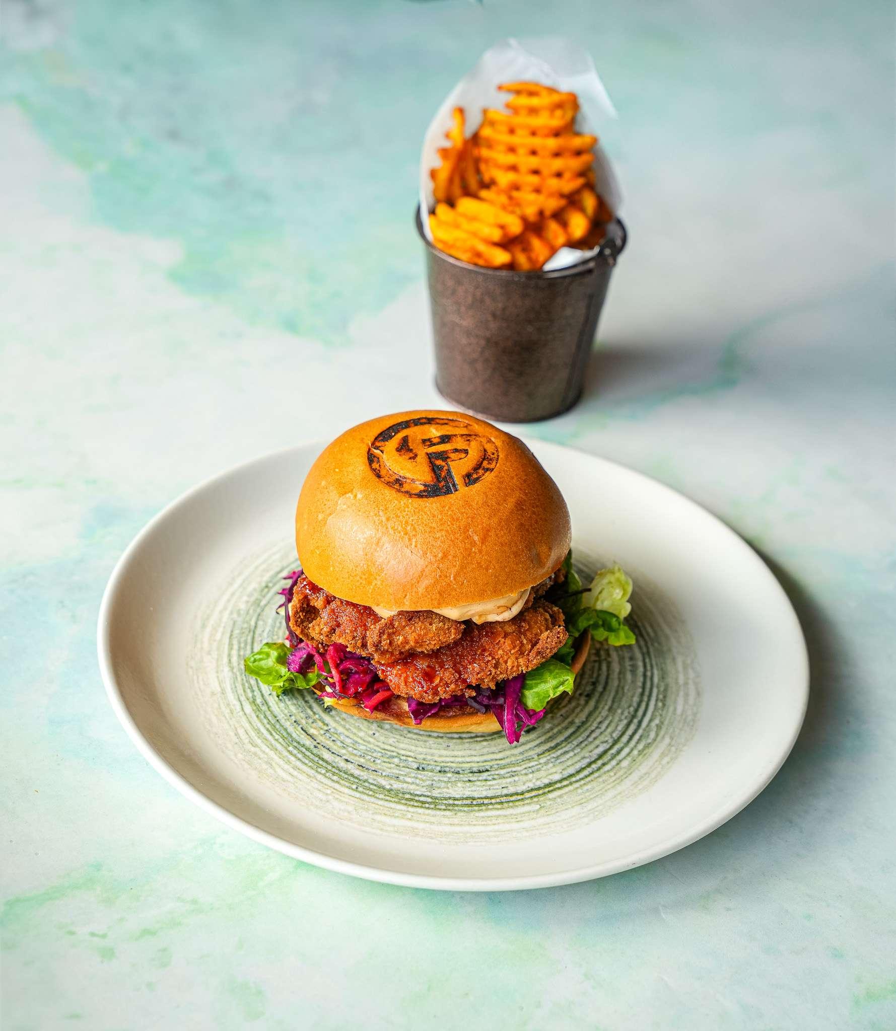 Crispy chicken burger with lettuce and slaw in a bun branded with a logo, served with waffle fries in a small metal bucket.