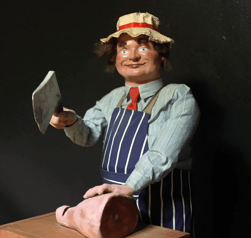 Lifelike mannequin of a smiling butcher wearing a striped apron, blue shirt, red tie, and straw hat with a red band, holding a meat cleaver above a large piece of ham on a wooden counter, displayed against a dark background.